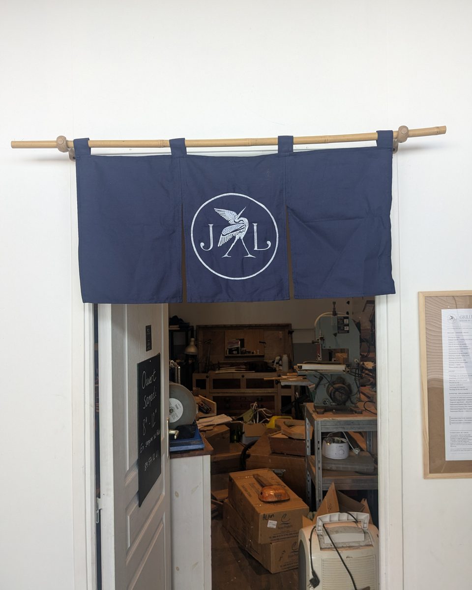 Porte de l'atelier à Fare Ute, sous un noren bleu brodé du héron JL